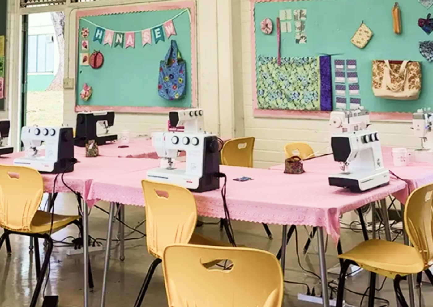Students at sewing machines in the Hana Sew studio with the Hana Sew banner on the wall
